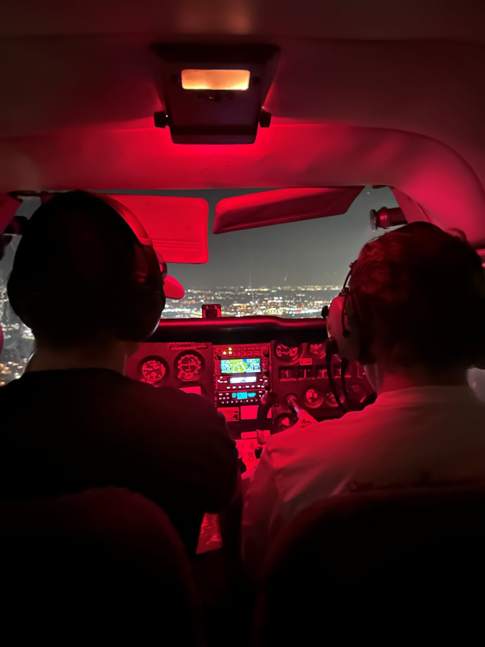 Two pilots flying at night under red cabin lighting, city lights visible below over the Dallas–Fort Worth area