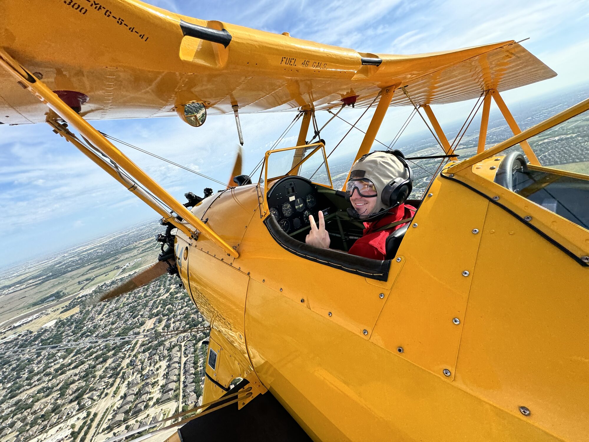 Reid Pekowski smiling and giving a peace sign from the open cockpit of a yellow biplane, flying over the Dallas–Fort Worth area